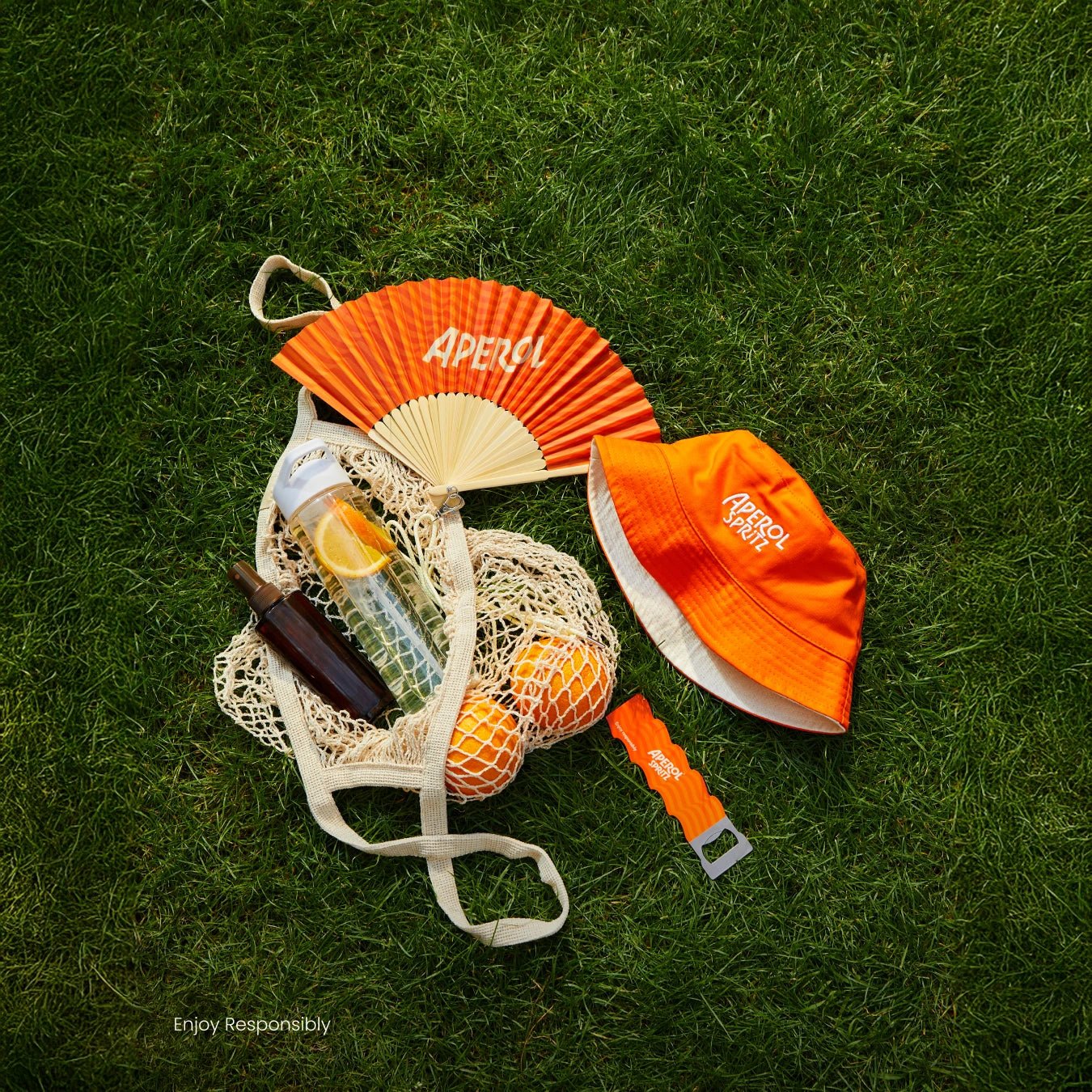 Aperol Spritz bucket hat, fan, bottle opener, and mesh bag with oranges on green grass